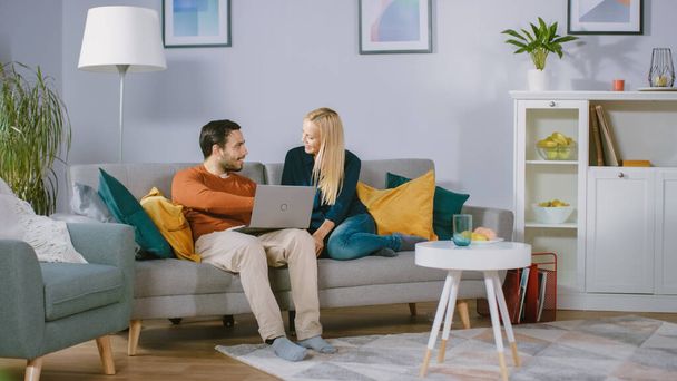 Happy Lovely Couple Sits on a Sofa at Home, Boyfriend Holds Laptop on Knees, Browsing through Internet, Doing e-Shopping, Using Streaming Services. Happy Family in Cozy Living Room. - Fotoğraf, Görsel