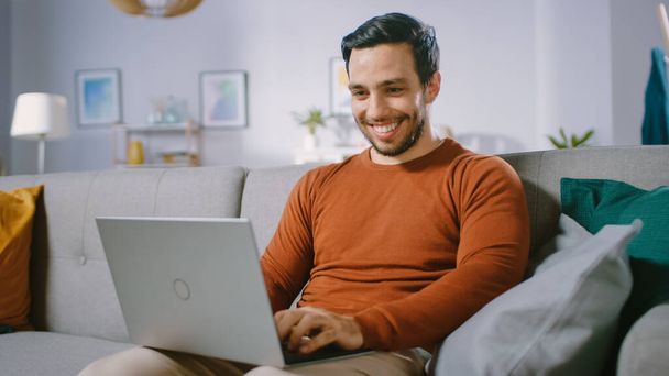 Cheerful Young Man at Home Sitting on a Sofa Holds Laptop on His Lap, Browses Through the Internet, Social Networks, Does e-Shopping. - 写真・画像