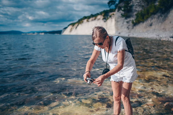 Yaşlı bir gezgin olgun bir kadın turist, deniz, taş, kaya ve mavi gökyüzünün arka planında fotoğraf çekiyor. Emekli insanlar yaz tatili, aktif yaşam tarzı konsepti - Fotoğraf, Görsel