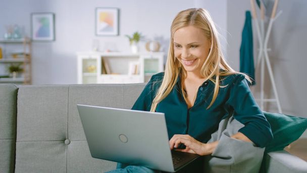 Schöne junge Frau entspannt sich zu Hause auf dem Sofa, nutzt Laptop, surft im Internet, schaut lustige Videos und macht E-Shopping. - Foto, Bild