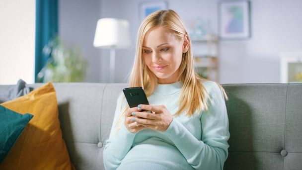 Beautiful Happy Pregnant Woman Sitting on a Couch Browses Through Internet on Smartphone. Future Mom Does Internet Shopping on Smartphone from Home. - Fotó, kép