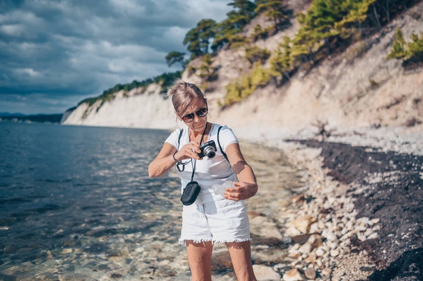 Idős idős utazó hátizsákos érett nő turista séta fotózás tengerparti háttér, kövek, sziklák, kék ég. Nyugdíjasok nyári vakáció, aktív életmód koncepció - Fotó, kép