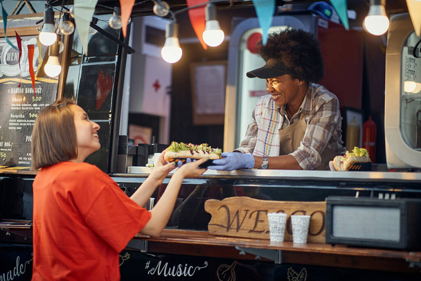 young caucasian satisfied customer taking order sandwiches from a cheerful afro-american employee in a modified truck for fast food service - Foto, imagen