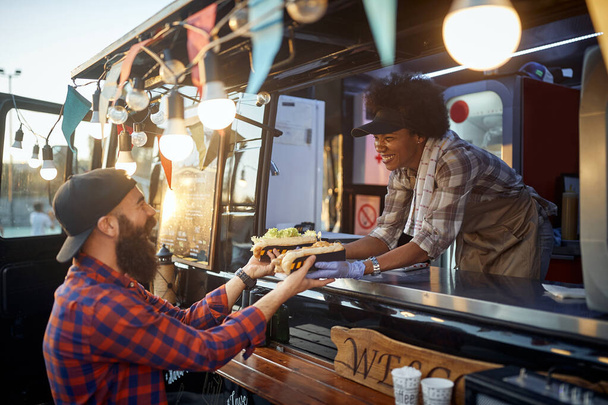young caucasian beardy hipster taking two sandwiches from polite female afro-american employee in fast food service - Fotografie, Obrázek