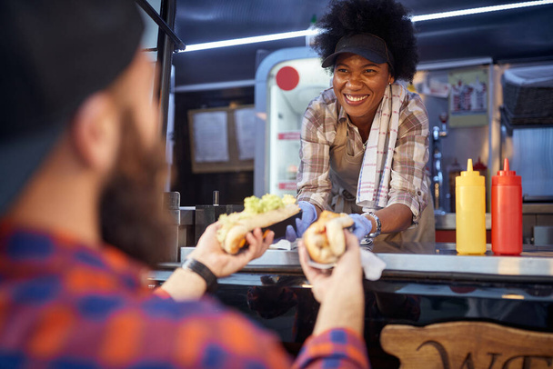 polite afro-american woman employee holding, giving, two sandwiches to a beardy caucasian male customer. fast food service - Zdjęcie, obraz