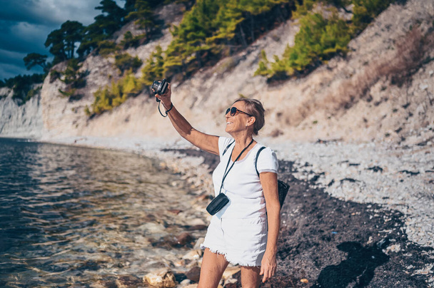 Idosos viajante sênior mochileiro mulher madura turista caminhando tirando fotos no fundo da costa do mar, pedras, pedras, céu azul. Pessoas aposentadas férias de verão, conceito de estilo de vida ativo - Foto, Imagem