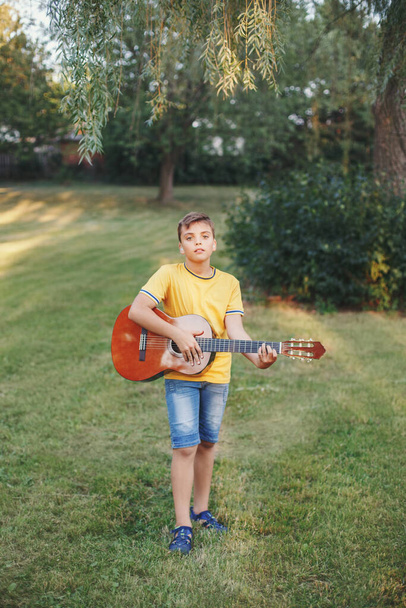 10代の少年が屋外でギターを弾くのを聞くのは難しい。公園で音楽や歌を演奏耳の補聴器を持つ子供。子供のためのホビーアート活動。本物の子供時代の瞬間.  - 写真・画像