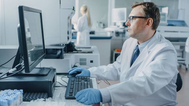 Male Research Scientist Sits at His Workplace in Laboratory, Uses Personal Computer. I the Background Genetics, Pharmaceutical Research Centre with Innovative Equipment. - Foto, immagini