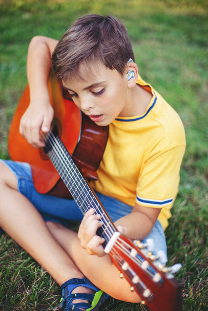Важко чути, як хлопчик грає на гітарі на відкритому повітрі. Дитина з слуховими апаратами в вухах грає музику і співає пісню в парку. Хобі художня діяльність для дітей. Справжній момент дитинства
.  - Фото, зображення