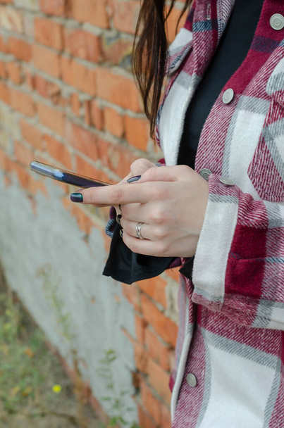 Les mains des femmes avec beau message de cadran de manucure dans le smartphone. Jeune femme en chemise à carreaux se tient avec téléphone au mur de briques. Mode de vie. - Photo, image