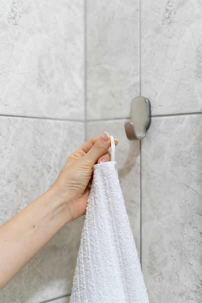 Vista recortada vertical da mão mulher pendura toalha de banho branco em um gancho no banheiro moderno com parede de espaço de cópia em azulejo - Foto, Imagem
