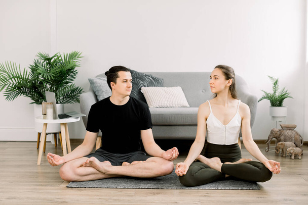 Young peaceful fit sporty couple, man and woman in sportswear, looking each other, meditating together sitting on floor mat, practicing pair yoga doing exercise in lotus pose at home - Foto, Imagem