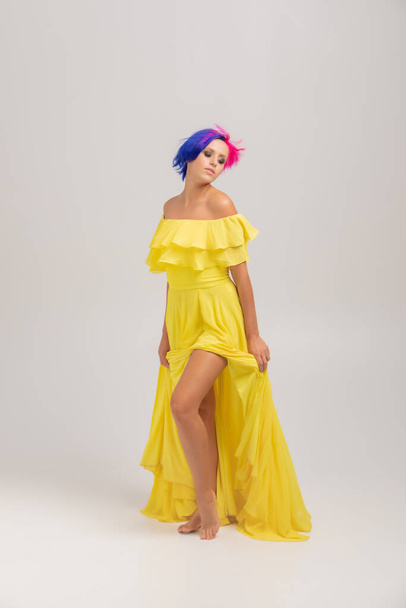 pełnowymiarowy portret kobiety z jasnymi kolorowymi włosami, niebieską i różową fryzurą. Farbowanie włosów, dziewczyna z krótkimi włosami na długie żółte sukienki z otwartymi ramionami stojących na lekkim cykloramie - Zdjęcie, obraz