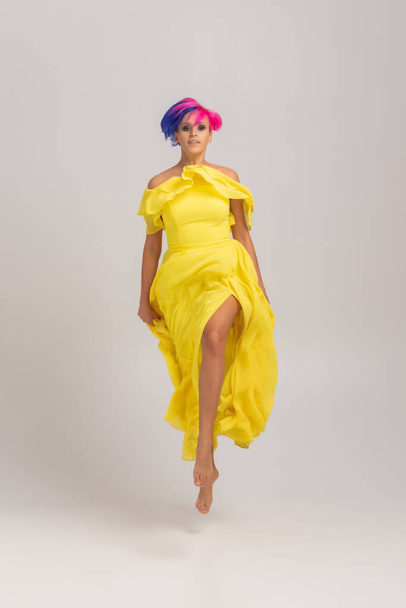 Ganzkörperporträt einer Frau mit hell gefärbten Haaren, blauem und rosa Haarschnitt. Haarfärbung, Mädchen mit kurzen Haaren trägt ein langes, gelbes Kleid mit offenen Schultern, das auf einem hellen Zyklorama springt - Foto, Bild
