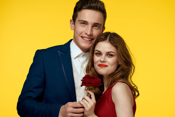 Um homem romântico abraça uma mulher em um vestido vermelho com uma rosa na mão em um fundo amarelo - Foto, Imagem