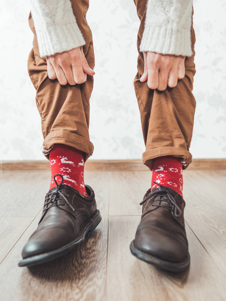Young man pulls up leg of his chinos trousers to show bright red socks with reindeers on them. Scandinavian pattern. Winter holiday spirit. Casual outfit for New Year and Christmas celebration. - Zdjęcie, obraz