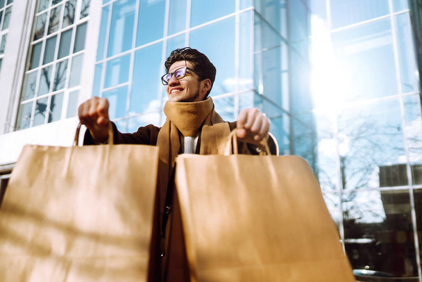 Young man holds out paper bags with purchases on the city street. Handsome man doing shopping in the city. Black friday. Autumn shopping.  - Foto, immagini