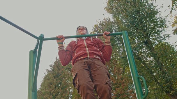 Młody nastolatek robi ćwiczenia ciągnąć na poziomym pasku podczas intensywnego treningu na świeżym powietrzu. Amerykański hipster trenuje samotnie w miejskim parku w wiosenny dzień. Osoba aktywna nosząca stylowe ubrania - Zdjęcie, obraz