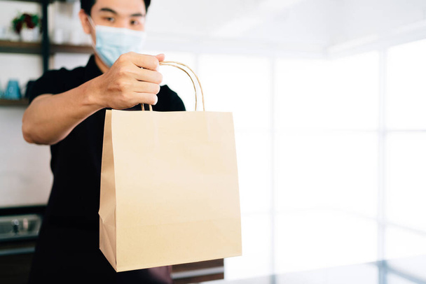 Male barista showing takeaway coffee in the paper bag during Coronavirus outbreak time. A worker inside the cafe counter for online delivery service. Coffee paper bag mockup. - Photo, Image
