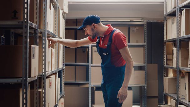 Handsome Warehouse Worker is Looking for a Cardboard Box on a Shelf with Other Packages. - Photo, image