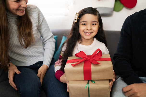 Piękna dziewczynka siedzi z rodzicami w czasie Bożego Narodzenia i trzyma kilka prezentów i wygląda bardzo szczęśliwy - Zdjęcie, obraz