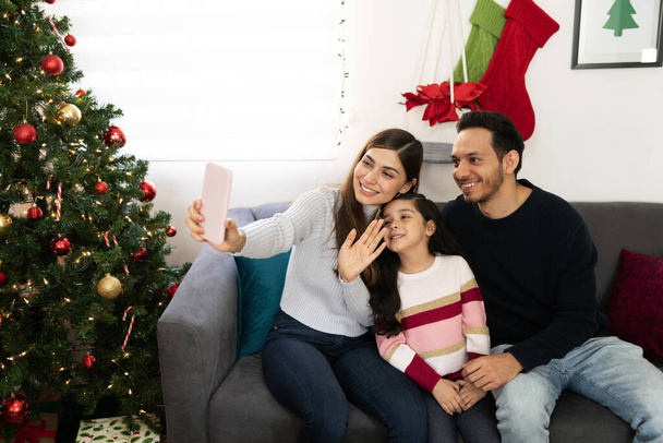 Όμορφη οικογένεια τριών ατόμων που χρησιμοποιούν smartphone για να κάνουν βιντεοκλήση με κάποιους συγγενείς τους τα Χριστούγεννα - Φωτογραφία, εικόνα