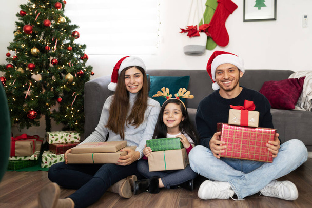 Feliz familia Hipanic sentada en el suelo en casa y sosteniendo muchos regalos de Navidad - Foto, Imagen