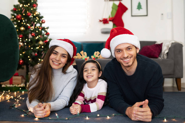 Porträt einer hispanischen dreiköpfigen Familie, die zu Hause neben einem Weihnachtsbaum liegt und bereit zum Feiern ist - Foto, Bild