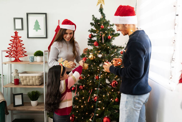 Linda niña ayudando a sus padres a poner algunos adornos y decoraciones en el árbol de Navidad en casa - Foto, Imagen