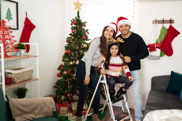 Retrato de uma bela família hispânica colocando luzes de corda e ornamentos na árvore de Natal juntos e sorrindo - Foto, Imagem