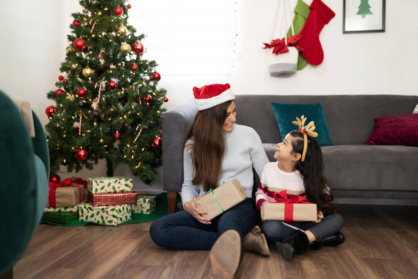 家で木の横に座っている間にクリスマスプレゼントを交換するかわいい母親と娘の肖像画 - 写真・画像