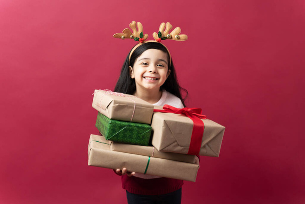 Πορτρέτο ενός όμορφου ισπανόφωνου μικρού κοριτσιού που κουβαλάει ένα μάτσο χριστουγεννιάτικα δώρα και δείχνει χαρούμενη - Φωτογραφία, εικόνα
