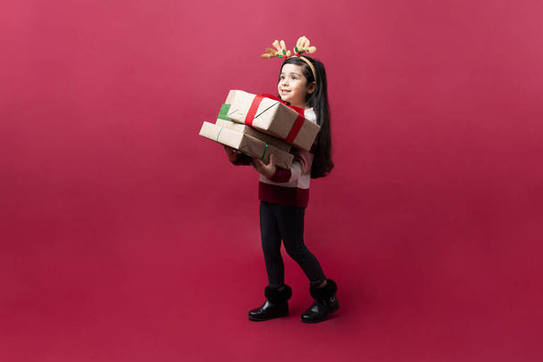 Ritratto a figura intera di una bambina che cammina con un mazzo di regali di Natale sullo sfondo rosso - Foto, immagini
