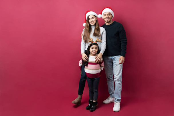 Vista completa de una feliz familia hispana lista para celebrar la Navidad y de pie sobre un fondo rojo - Foto, Imagen