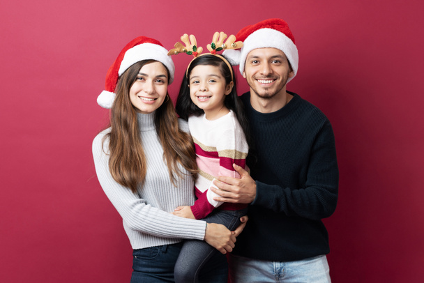 Familia hispana de tres abrazándose unos a otros de pie sobre un fondo rojo en Navidad - Foto, Imagen