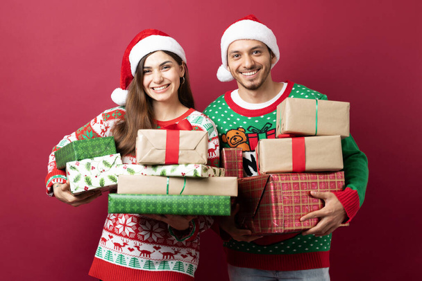 Schattig Latijns paar met een bos van kerstcadeaus en glimlachen tegen een rode achtergrond in een studio - Foto, afbeelding