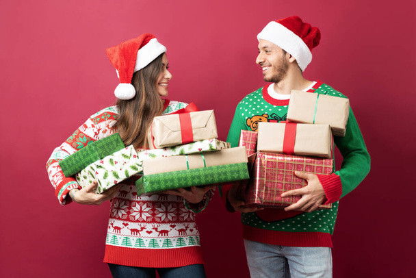 ハッピーヒスパニックカップル身に着けているサンタの帽子と運ぶ多くのクリスマスプレゼントでスタジオ - 写真・画像