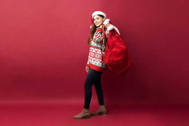 Ganzkörperansicht einer brünetten Frau mit Weihnachtsmütze, die eine Tasche trägt und ein paar Weihnachtsgeschenke ausliefert - Foto, Bild