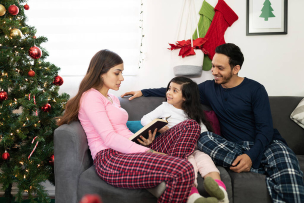 Jolie famille latine assise près du sapin de Noël portant un pyjama et lisant quelques histoires ensemble - Photo, image
