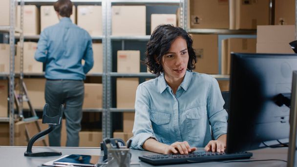 Female Inventory Manager Sitting at Her Desk and Using Personal Computer, Worker Puts Package on the Designated Shelf. In the Background Rows of Cardboard Boxes with Products Ready For Shipment. - 写真・画像