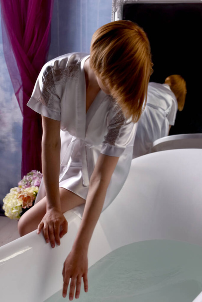 Frau ohne Gesicht im Bademantel sitzt am Rand der Badewanne und füllt sich mit Wasser - Foto, Bild