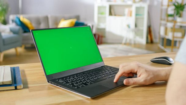 Man Uses Laptop with Green Mock-up Screen While Sitting at the Desk in His Cozy Living Room. - Foto, Imagem