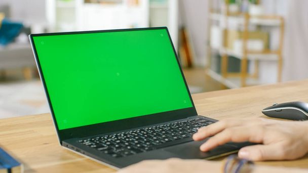 Homem usa laptop com tela de Mock-up verde enquanto sentado na mesa em sua acolhedora sala de estar. - Foto, Imagem