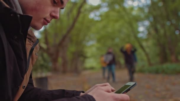 Fiatal fickó telefonnal a kezében ül egy padon a parkban. A háttérben egy baráti társaság sétál a parkban. A magány és az internettől és okostelefonoktól való függés fogalma. - Felvétel, videó