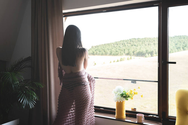Sensual woman wrapped in a shawl admiring view from house window - Foto, afbeelding