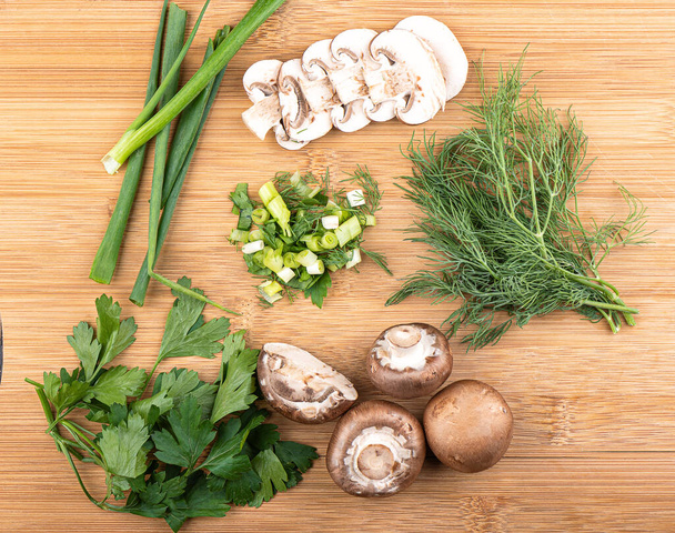 Top view. Sliced and whole mushrooms, green onions, parsley and dill on a wooden background. Chopped greens in the center. Close up. - Фото, изображение