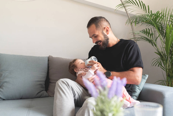 Loving father feeding bottled milk to baby daughter sitting on sofa at home - Zdjęcie, obraz