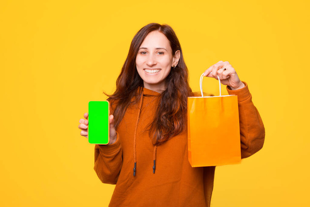 Een vrolijke jonge vrouw houdt een boodschappentas en een telefoon vast. - Foto, afbeelding