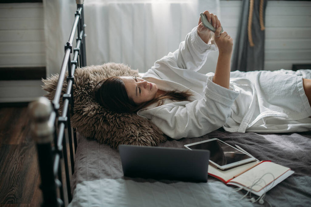 Een vrouw rust op een bed in een slaapkamer met een telefoon. Een jong meisje ligt thuis op een groot bed, communiceert online, neemt een selfie. Rustpauze - Foto, afbeelding