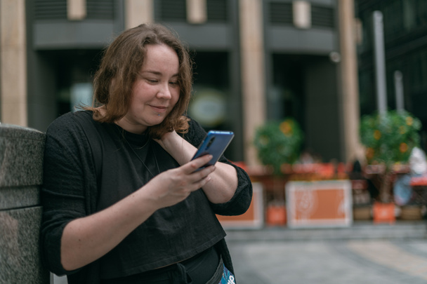 Portrait of a young woman with a phone in a big city. A chubby beautiful girl stands with a smartphone in her hands and communicates on the Internet on the street of a modern metropolis - Fotó, kép